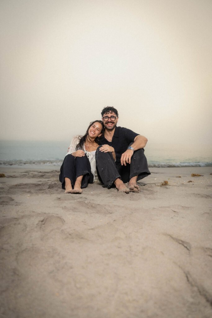 Casual couple sitting together on sand West Palm Beach engagement photography relaxed pose