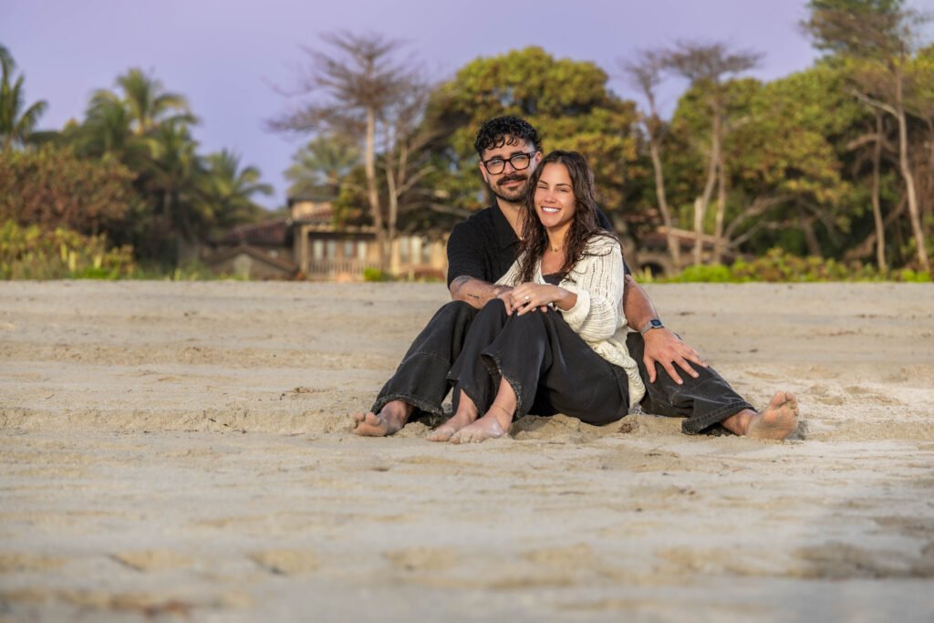 Engaged couple sitting barefoot on the sand at a Palm Beach beach, smiling at the camera