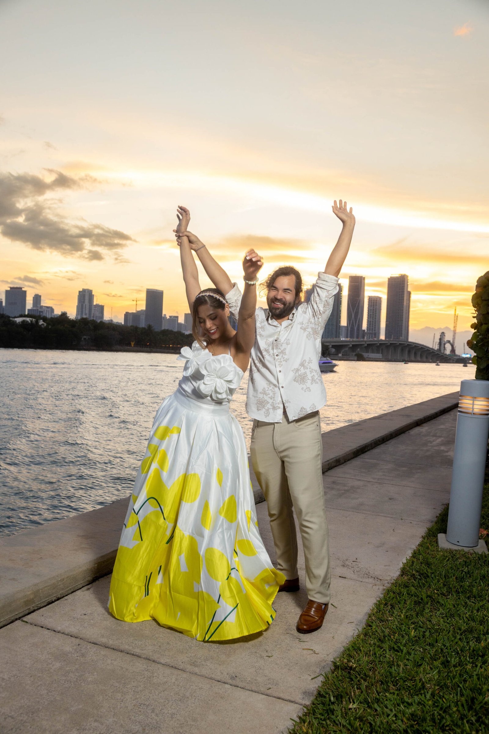 Joyful engaged couple celebrating with arms raised at sunset near the West Palm Beach waterfront by The Love Pics