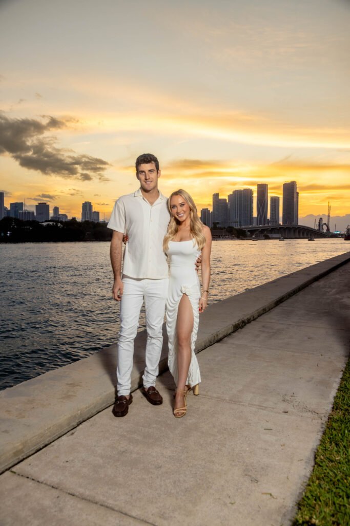 Couple in white outfits posing together at a waterfront promenade during a sunset engagement portrait session