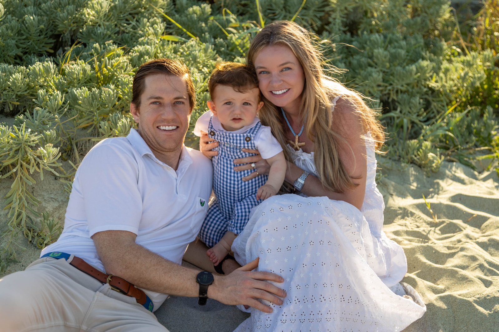Smiling young family with their toddler son posing in the sand at a beach session with dunes and vegetation background