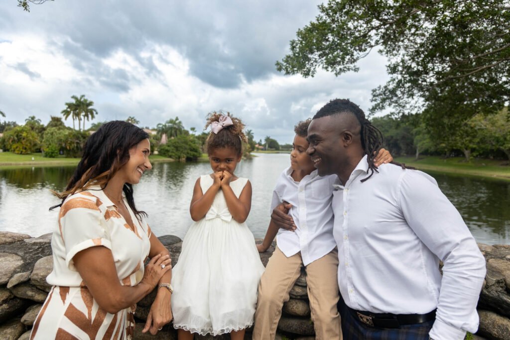 Smiling family of four with two young daughters posing together at a lake with tropical trees background during an outdoor portrait session