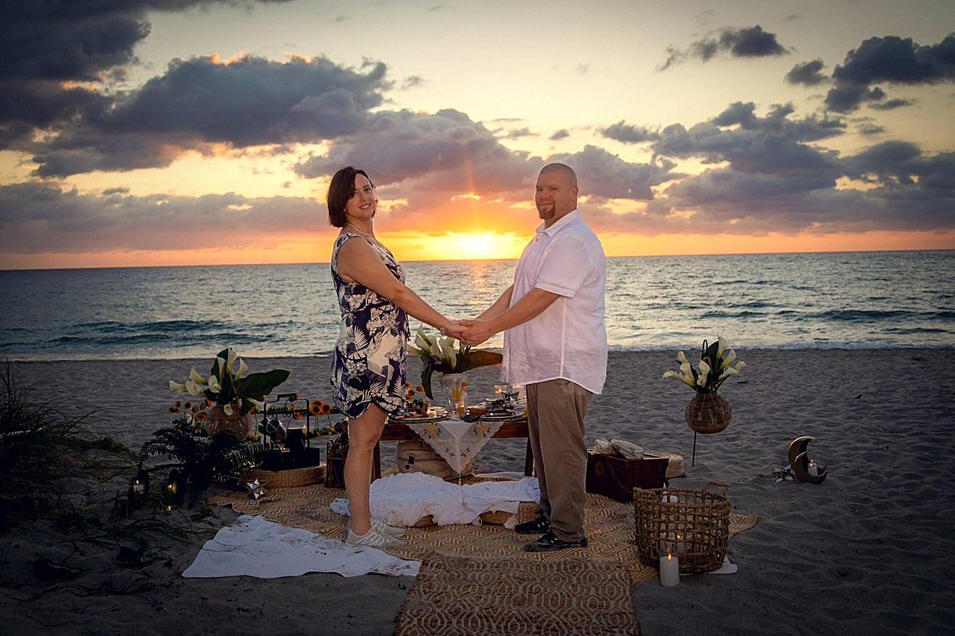 Engaged couple holding hands with the sunrise between them above a flower-and-candle beach proposal setup, Fort Lauderdale