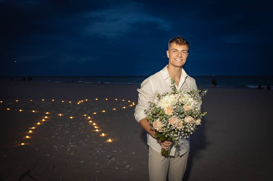 Newly engaged man smiling with a bridal bouquet and a candlelit beach behind him in West Palm Beach