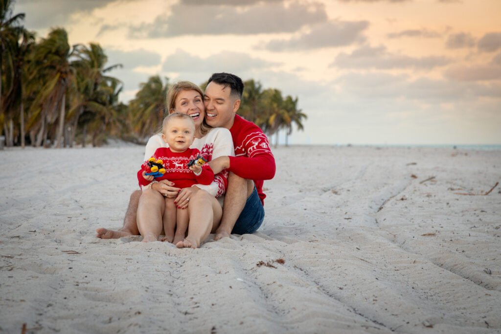 Smiling family portrait at the beach with parents and son sitting in the sand in Christmas sweaters during sunset session