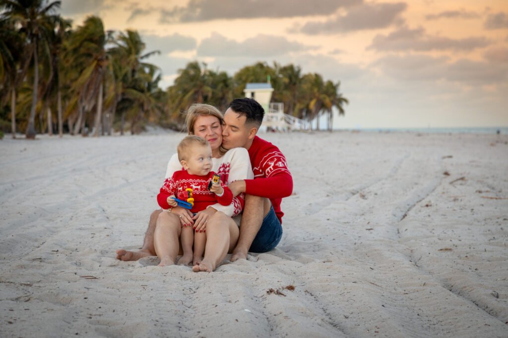 Loving family portrait at the beach with parents kissing their son in red Christmas sweaters during sunset session