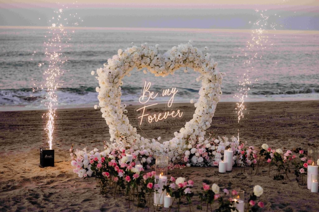 Heart-shaped rose arch with Be My Forever neon sign, cold-spark fountains and candles on the beach at dusk — luxury proposal setup by The Love Pics