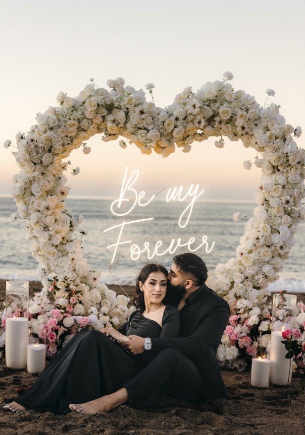 Couple embracing in front of a floral heart arch with neon sign during a beach proposal by The Love Pics