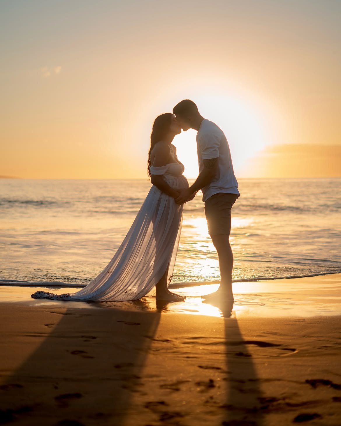 Expecting mother in a flowing white dress on the beach at golden hour