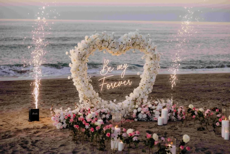 Heart-shaped rose arch with Be My Forever neon sign, cold-spark fountains and candles on the beach at dusk — luxury proposal setup by The Love Pics
