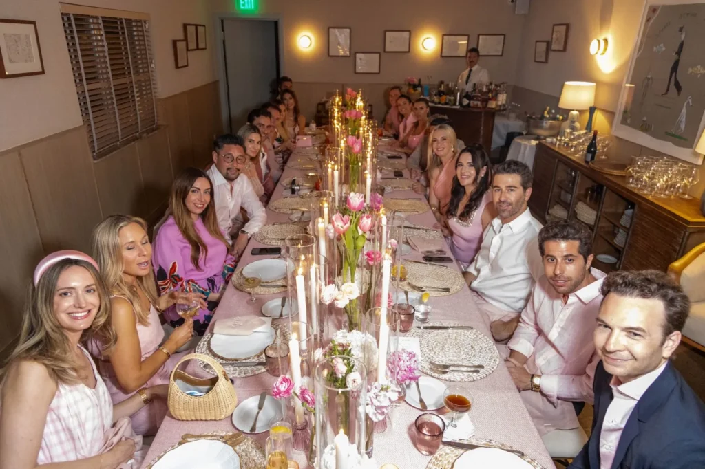 Elegant group of friends in pink outfits at a long dinner table with floral centerpieces and candles at an indoor event