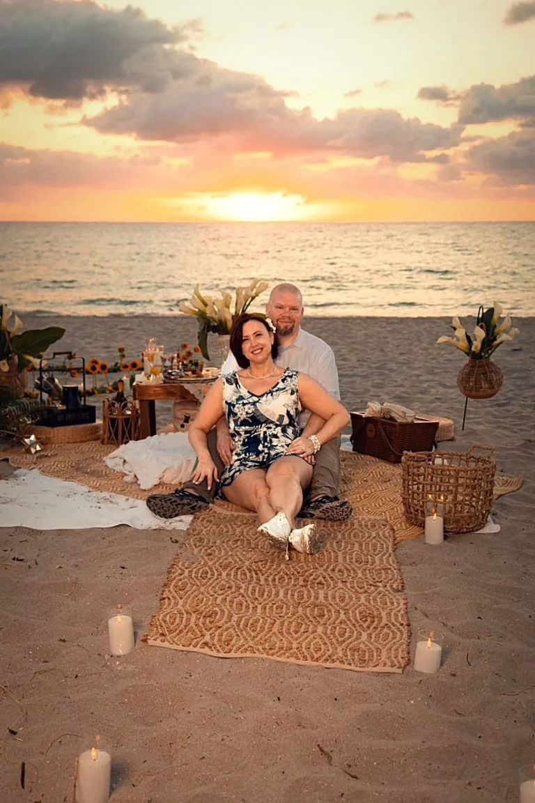 Couple at a luxury boho beach picnic proposal setup with sunflowers, calla lilies and candles during a golden sunrise at Las Olas Beach, Fort Lauderdale, by The Love Pics.