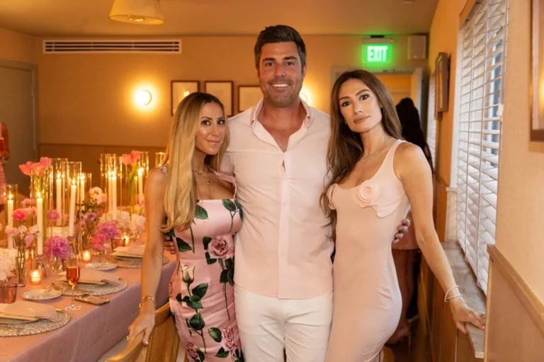 Man posing with two elegant women in pink dresses at an indoor event with floral centerpieces and candles