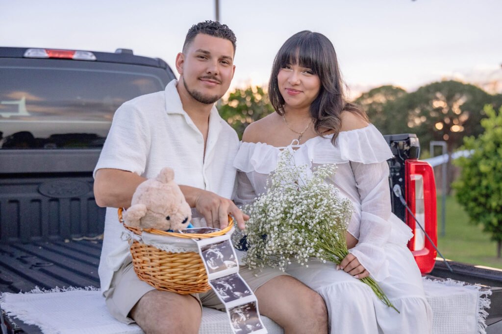 Expecting couple dressed in white sitting on a pickup tailgate at sunset with a teddy bear basket and baby's breath