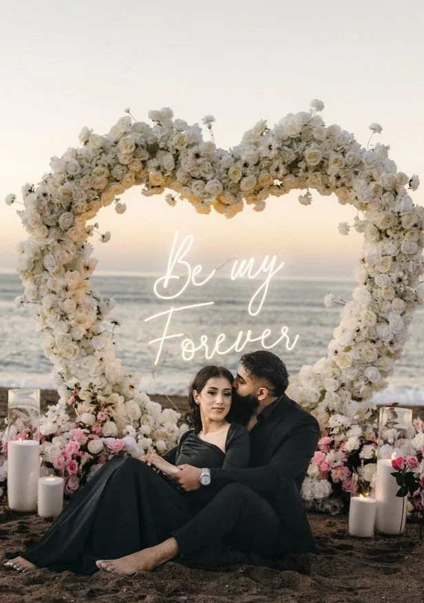 Pareja abrazándose frente a un arco floral de corazón en una propuesta en la playa