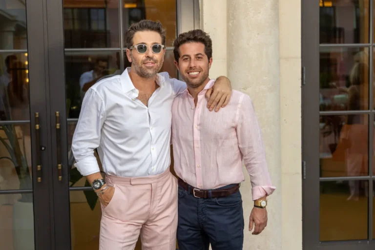 Two elegant men in pink and white shirts smiling at an indoor event with architectural background