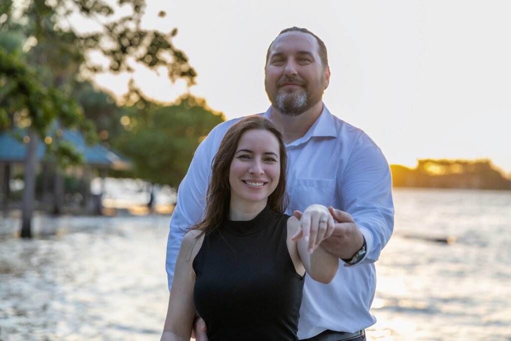The couple smiles at the camera by the water at sunset while she shows her engagement ring