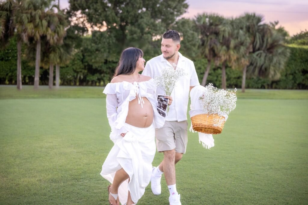 Expecting couple in white walking across a lawn with a basket and an ultrasound print, Royal Palm Beach