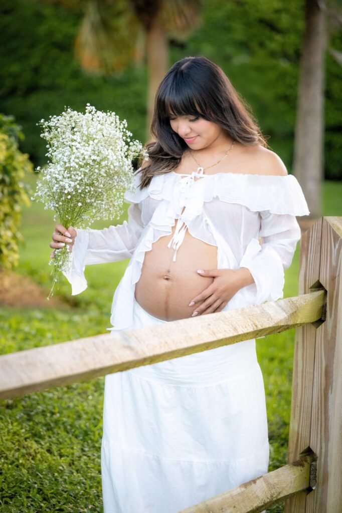 Pregnant woman in white holding baby's breath beside a wooden fence in a green park, Royal Palm Beach