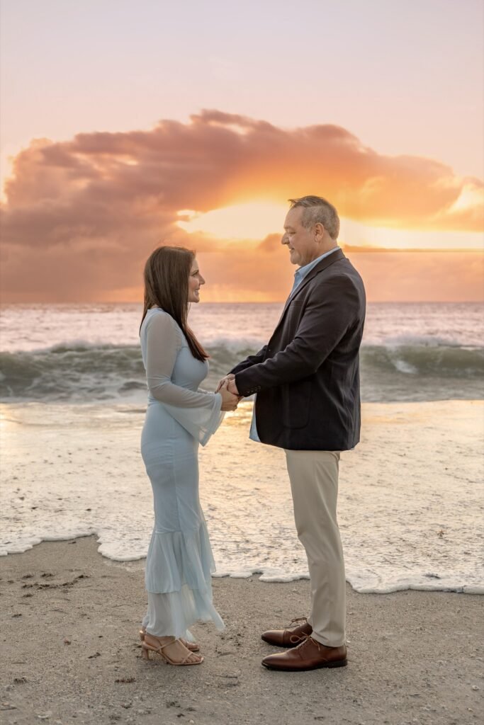 Couple in pale blue and navy holding hands at the shoreline under a sunset sky